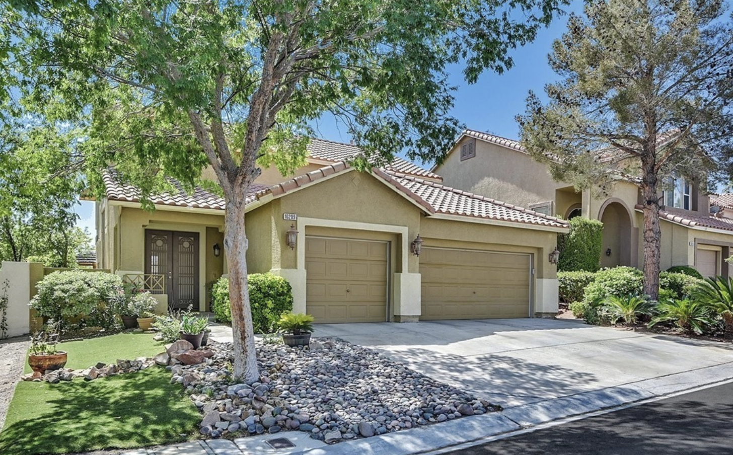 A beautifully landscaped beige home full-sized trees, turf, bushes, palms, and stone garden beds